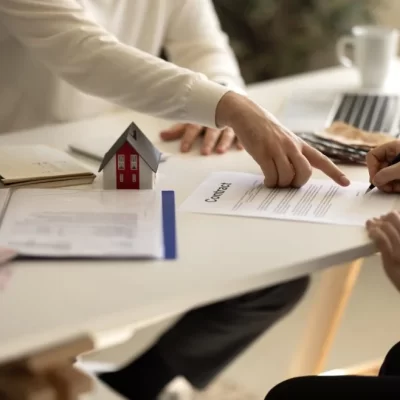 A cena retratada é de uma reunião em um escritório, onde duas pessoas discutem um contrato, com uma delas prestes a assinar. A mesa contém diversos documentos, um modelo de casa, e um laptop, indicando que a conversa gira em torno de um acordo relacionado à compra ou venda de um imóvel para a lei do inquilinato em um conteudo da group software