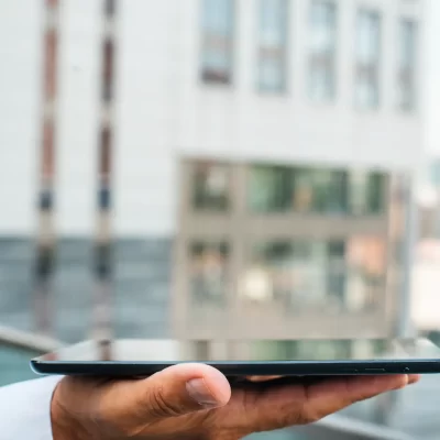 Uma mão segura um tablet em frente a um condomínio inteligente.