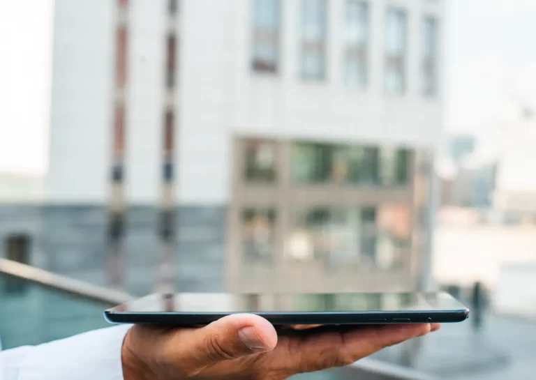 Uma mão segura um tablet em frente a um condomínio inteligente.