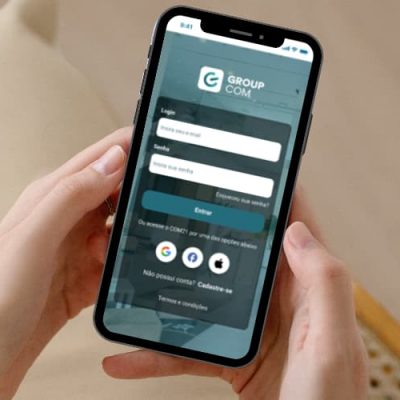 Imagem de duas mãos segurando um celular com a tela de login do GroupCOM.