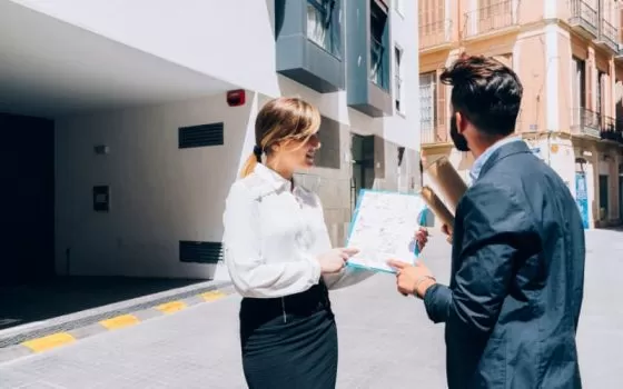 Imagem de uma mulher loira com roupa social apresentando um documento em uma prancheta para um homem de terno. O homem olha para um prédio em segundo plano, ilustrando o processo de locação comercial.