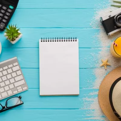 Imagem de uma mesa de trabalho com objetos relacionados à gestão de férias. A imagem inclui um notebook, um teclado, uma xícara de café e uma calculadora do lado esquerdo, um bloco de notas no centro e areia, uma câmera fotográfica, um chapéu de praia e um um copo de suco de laranja do lado direito.