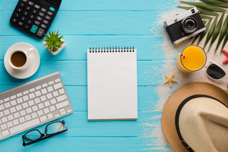 Imagem de uma mesa de trabalho com objetos relacionados à gestão de férias. A imagem inclui um notebook, um teclado, uma xícara de café e uma calculadora do lado esquerdo, um bloco de notas no centro e areia, uma câmera fotográfica, um chapéu de praia e um um copo de suco de laranja do lado direito.