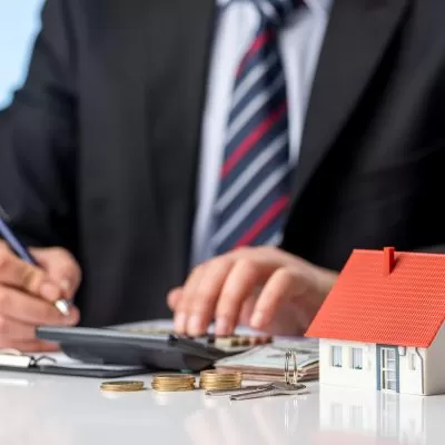 Homem de negócios em terno calculando finanças com uma calculadora, representando a administração financeira de um condomínio. Na mesa, há uma pequena maquete de uma casa, chaves e moedas empilhadas. Qual o índice para reajuste de taxa de condomínio? A cena simboliza a importância de determinar corretamente os índices de reajuste das taxas condominiais.