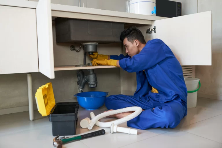 Um homem vestindo um macacão azul e luvas amarelas, trabalhando para limpar e desentupir caixa de gordura em uma pia na cozinha. Ele está sentado no chão, com suas ferramentas ao seu lado.