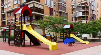Playground em uma área de lazer de condomínio, com dois escorregadores amarelos, uma área de escalada com pedras e brinquedos de mola em forma de elefante, cercado por edifícios residenciais e árvores.