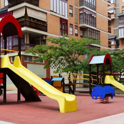 Playground em uma área de lazer de condomínio, com dois escorregadores amarelos, uma área de escalada com pedras e brinquedos de mola em forma de elefante, cercado por edifícios residenciais e árvores.