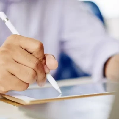 A imagem retrata uma mão segurando uma caneta stylus, escrevendo em um tablet, simbolizando a assinatura digital de documentos. O ambiente é de trabalho, com um fundo desfocado que sugere concentração e produtividade.