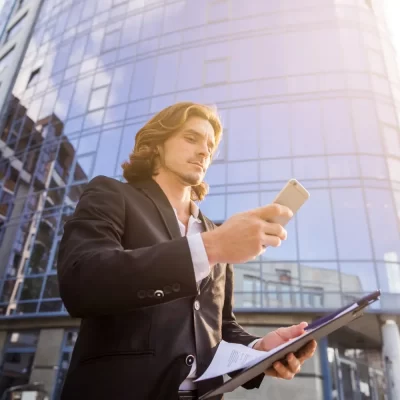 Um homem com terno escuro está em frente a um edifício moderno, segurando uma pasta e usando um smartphone, de forma a representar a automação de tarefas do síndico.