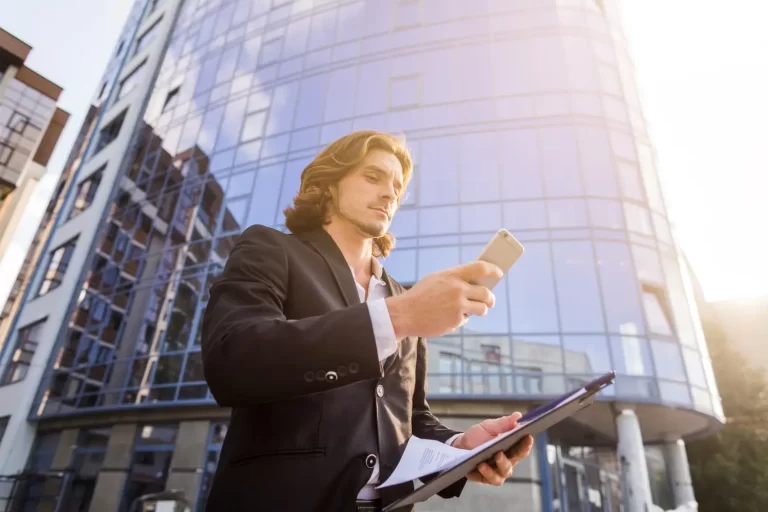 Um homem com terno escuro está em frente a um edifício moderno, segurando uma pasta e usando um smartphone, de forma a representar a automação de tarefas do síndico.