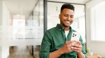 O homem sorridente, usando uma camisa verde, está concentrado em seu smartphone em um ambiente de escritório moderno. Ele parece satisfeito, possivelmente utilizando um aplicativo de vistoria por voz para fazer um laudo técnico