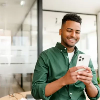 O homem sorridente, usando uma camisa verde, está concentrado em seu smartphone em um ambiente de escritório moderno. Ele parece satisfeito, possivelmente utilizando um aplicativo de vistoria por voz para fazer um laudo técnico