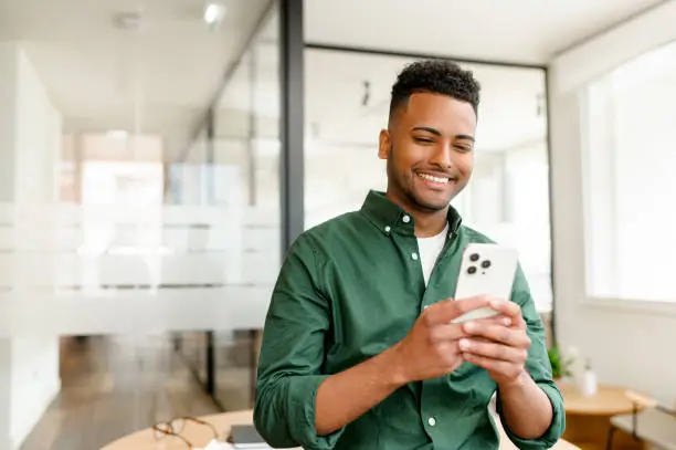 O homem sorridente, usando uma camisa verde, está concentrado em seu smartphone em um ambiente de escritório moderno. Ele parece satisfeito, possivelmente utilizando um aplicativo de vistoria por voz para fazer um laudo técnico