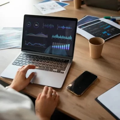 Uma pessoa elabora um planejamento orçamentário condominial, usando um laptop com gráficos na tela e rodeada de documentos impressos em uma mesa de trabalho.