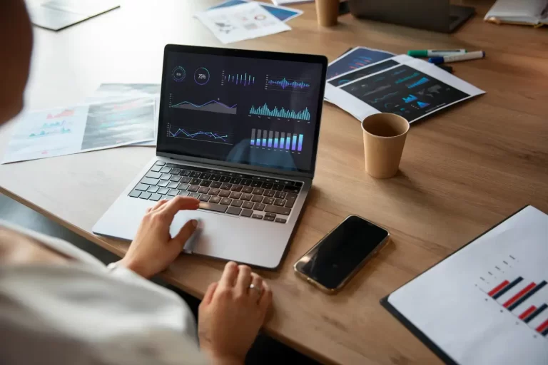 Uma pessoa elabora um planejamento orçamentário condominial, usando um laptop com gráficos na tela e rodeada de documentos impressos em uma mesa de trabalho.